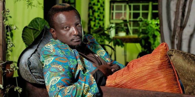 This is a photograph of Binyavanga Wainaina leaning back on a cushioned chair, wearing a colouful patterned jacket and looking at the camera.