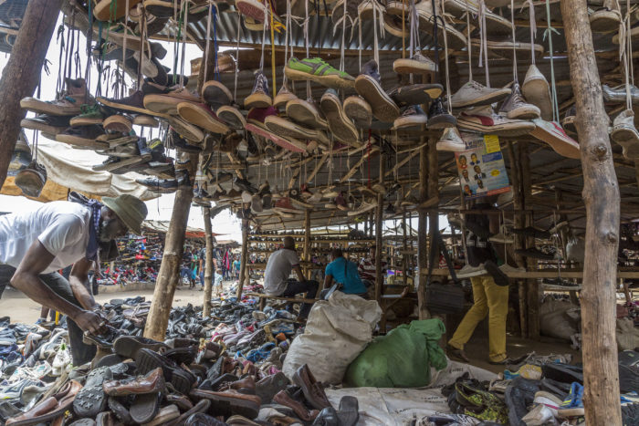 This photograph shows one of the covered stalls selling shoes. There is a man on the left searching through the shoes on the ground. there are shoes hanging by their laces from the roof of the staff. there are other stalls and traders in the background.