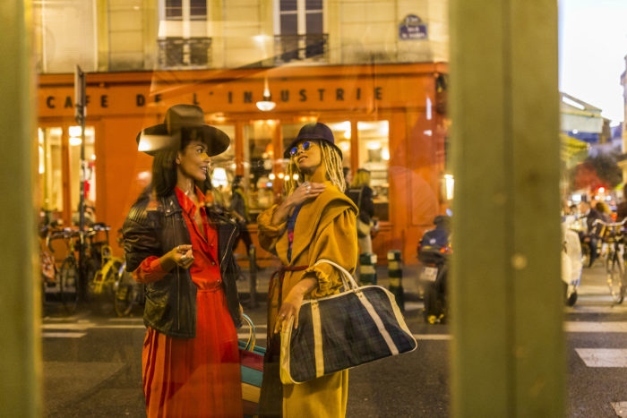 This is a photograph of two women taken from inside a shop through the window. The woman on the left is wearing a cowboy hat, black jacket and red dress. and is holding a colourful stiped bag. The woman on the right is wearing a black hat, reflective sunglasses and an yellow/orange coat and is carrying a canvas bag. There is a café across the street in the background and a level crossing to the right of the image.