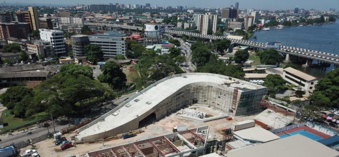 The image shows the John K Randle Heritage Centre under construction. The city of Lagos is in the background with the Lagos Lagoon to the right of the Heritage Centre.