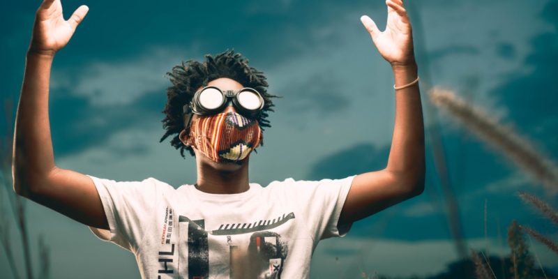 Man wearing patterned fabric face mask and goggles, stood with his arms raised in front of a grey, cloudy sky