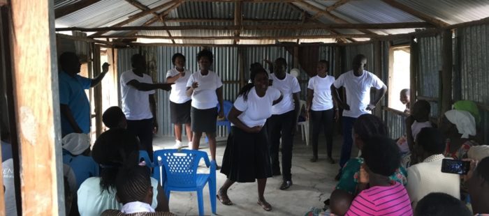 This is an image of a group of young people wearing white t-shirts and black trousers / skirts performing in front of a seated audience in corrugated iron building.